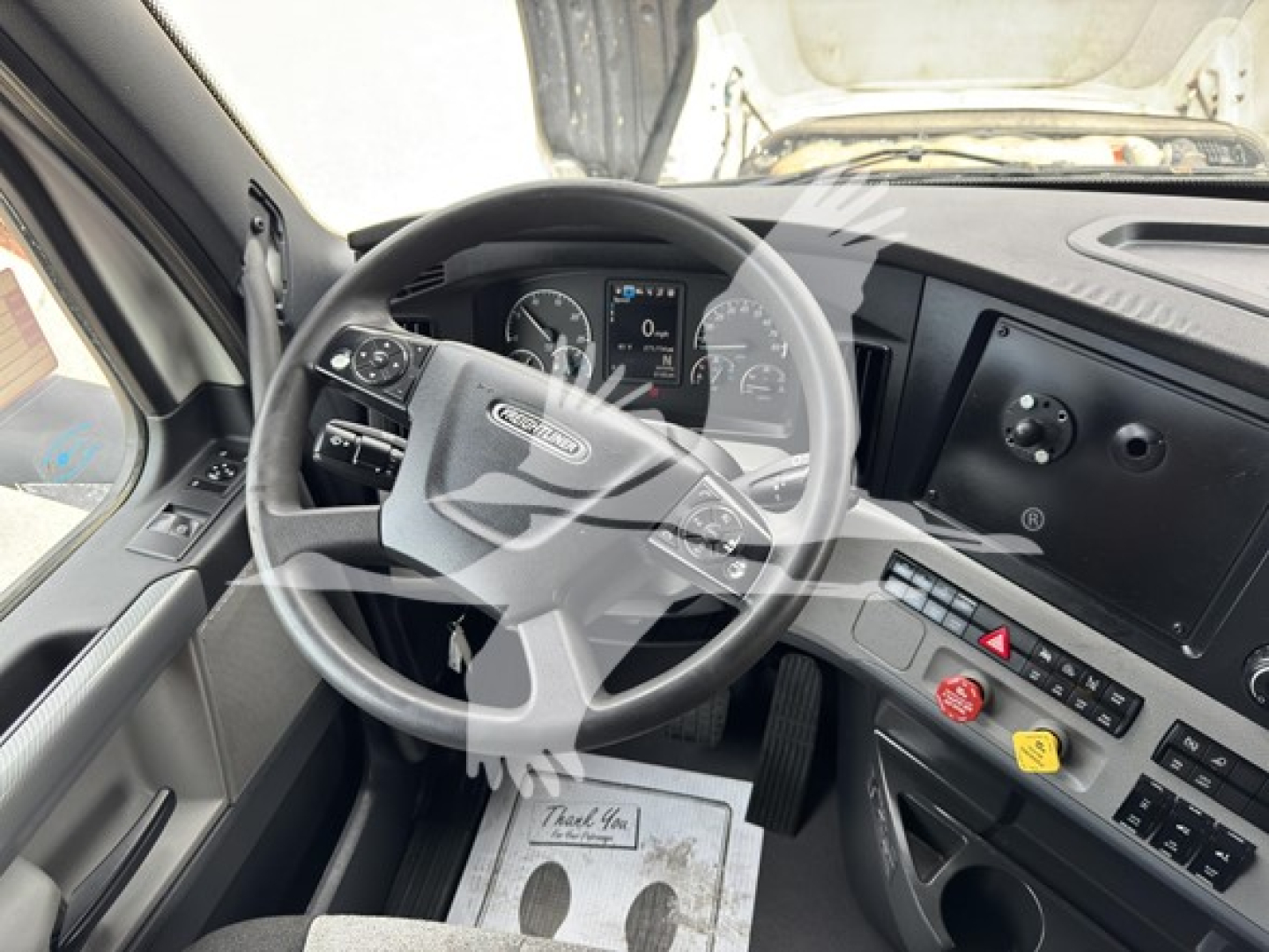 Freightliner Cascadia truck interior showing steering wheel with controls, dashboard with digital gauge cluster, center console with hazard button, climate and utility switches, and two colored knobs.