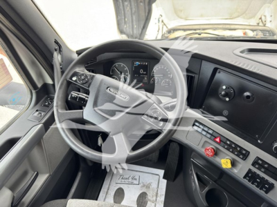 Freightliner Cascadia truck interior showing steering wheel with controls, dashboard with digital gauge cluster, center console with hazard button, climate and utility switches, and two colored knobs.