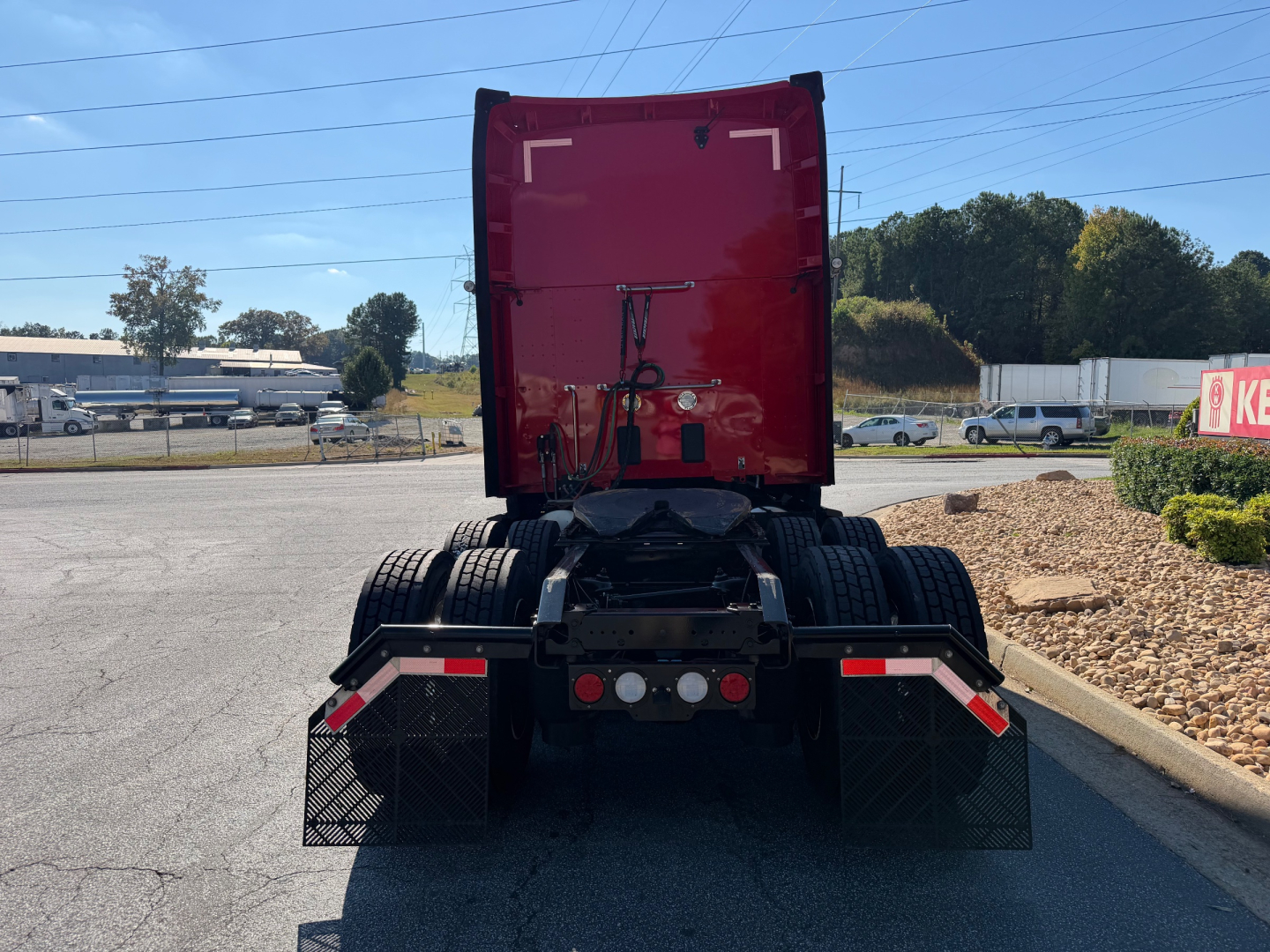 Kenworth T680 rear view showing fifth wheel and dual rear axles with mud flaps and red sleeper cab in background.