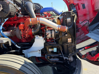 Kenworth T680 engine bay showing red engine block, silver alternator, blue intercooler piping, orange turbocharger intake, white coolant reservoir, and black radiator support.