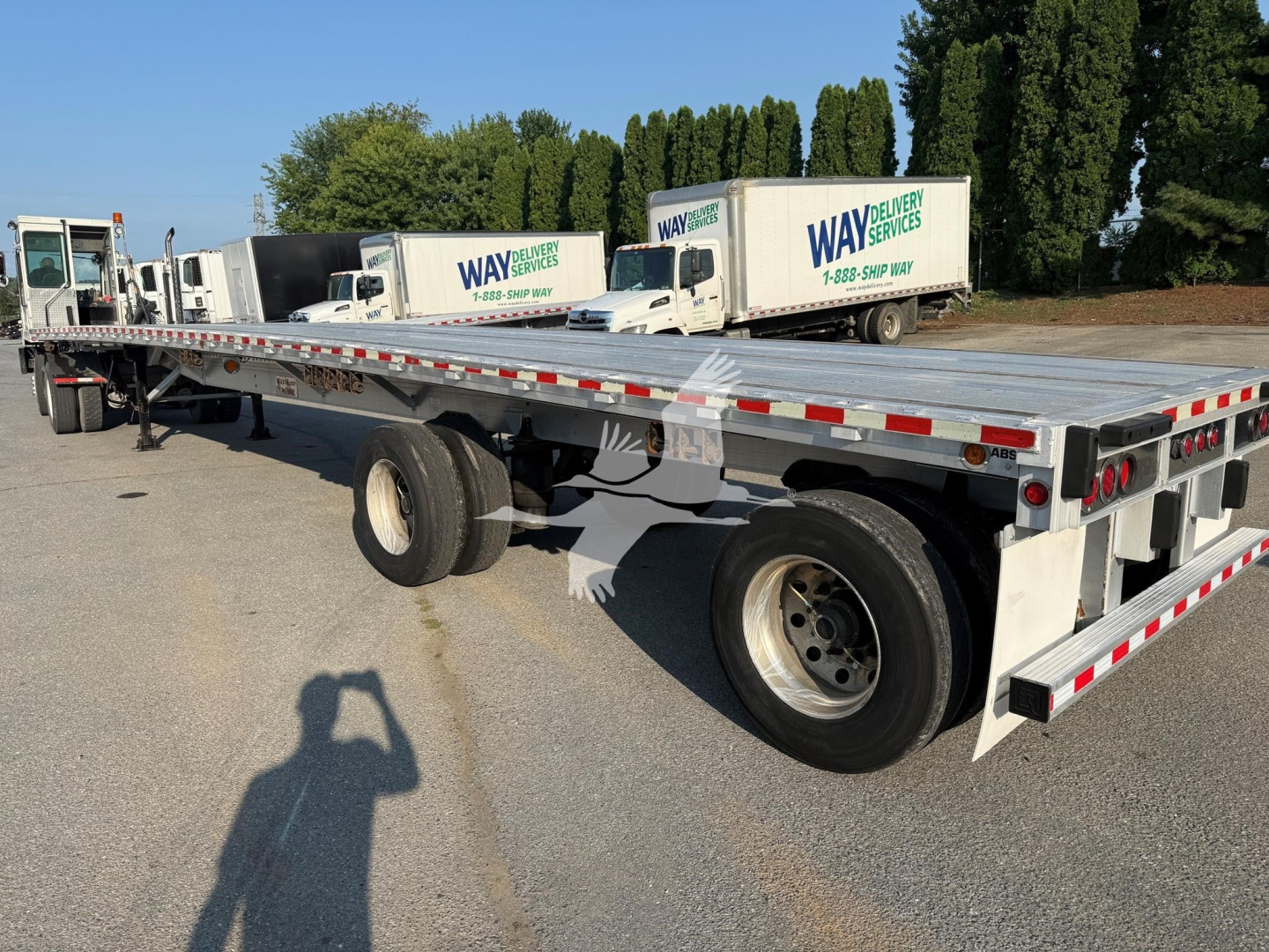 Flatbed trailer showing long aluminum deck and tandem axles with Way Delivery Services trucks in background.