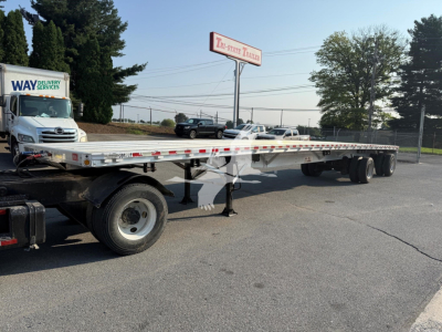 Flatbed trailer in a dealership lot showing long aluminum deck and dual axles with ramps and support legs.