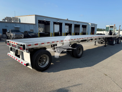 Flatbed trailer showing long aluminum deck and dual rear axles with wheel hubs, taillights, and reflective tape; truck yard and service bays in background.