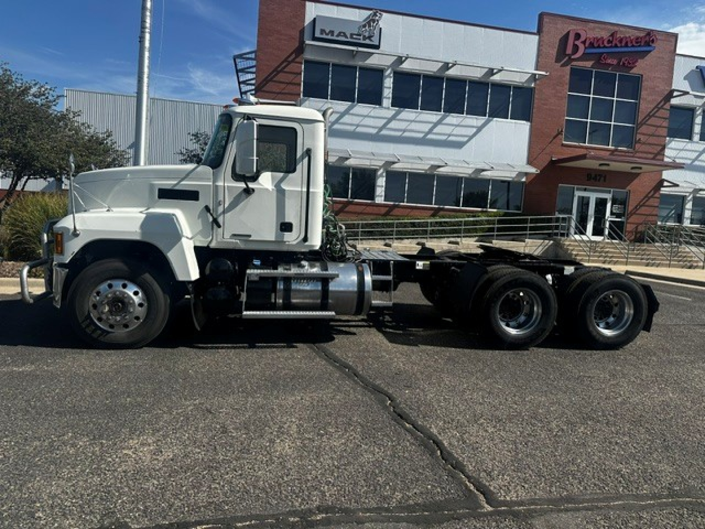 Truck side view showing 2020 Mack PI64T conventional daycab with three rear axles and flatbed frame in a dealership lot.