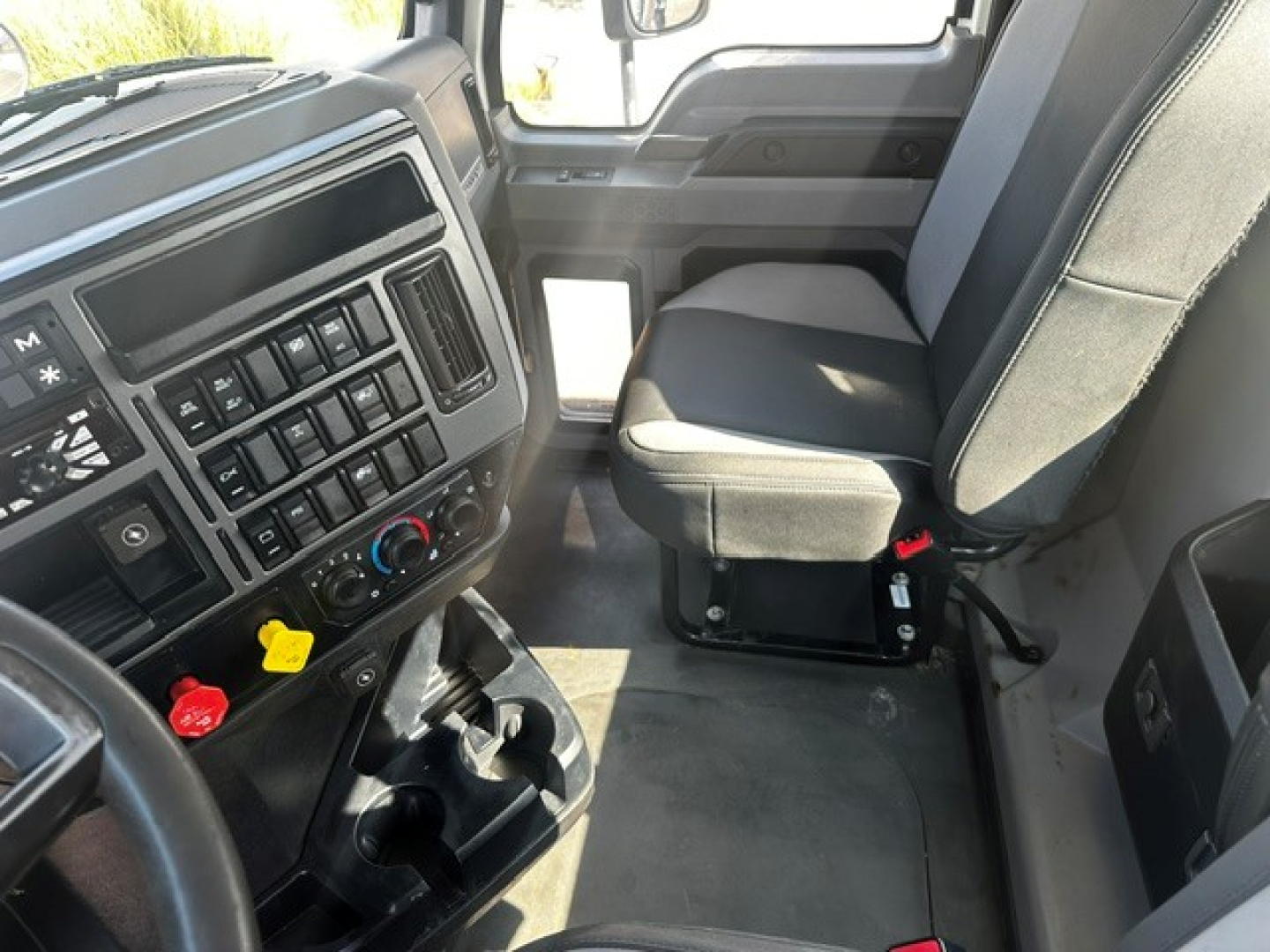 Truck interior showing dash by steering wheel with control buttons, climate controls, cup holders, and driver seat in a Mack PI64T cab.