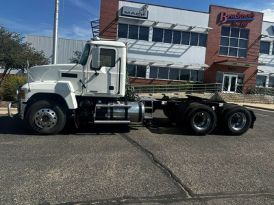 Truck side view showing 2020 Mack PI64T conventional daycab with three rear axles and flatbed frame in a dealership lot.