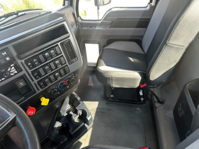 Truck interior showing dash by steering wheel with control buttons, climate controls, cup holders, and driver seat in a Mack PI64T cab.