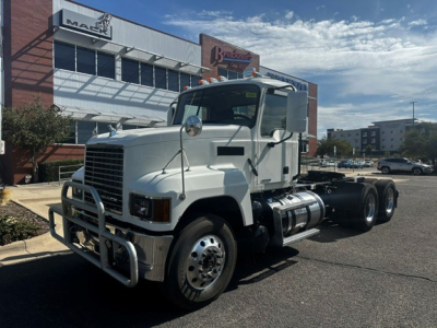 White Mack PI64T conventional day cab truck in front of a dealership, showing the front bumper guard and passenger side; no visible text on the truck itself.