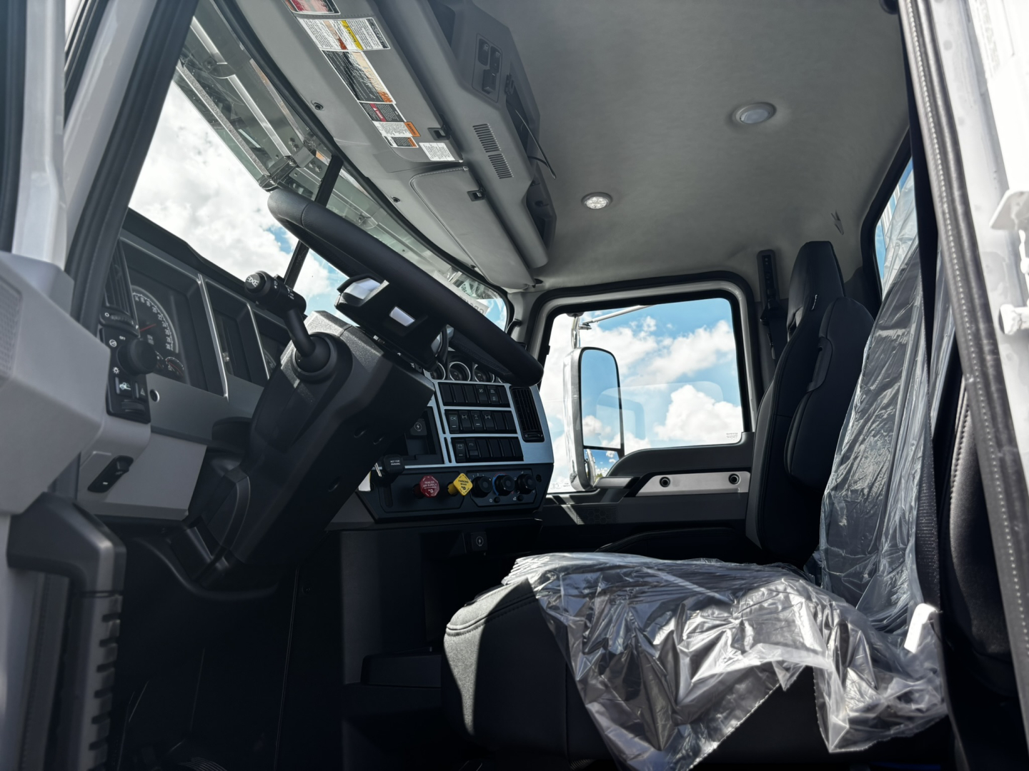Truck cab interior of a 2027 Mack GR64FT showing steering wheel, dashboard, and driver seat with protective plastic.