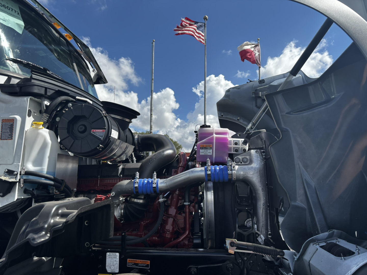 Front engine bay of a 2027 Mack GR64FT conventional daycab showing engine components, hoses, reservoir with yellow cap, radiator, and purple coolant reservoir.