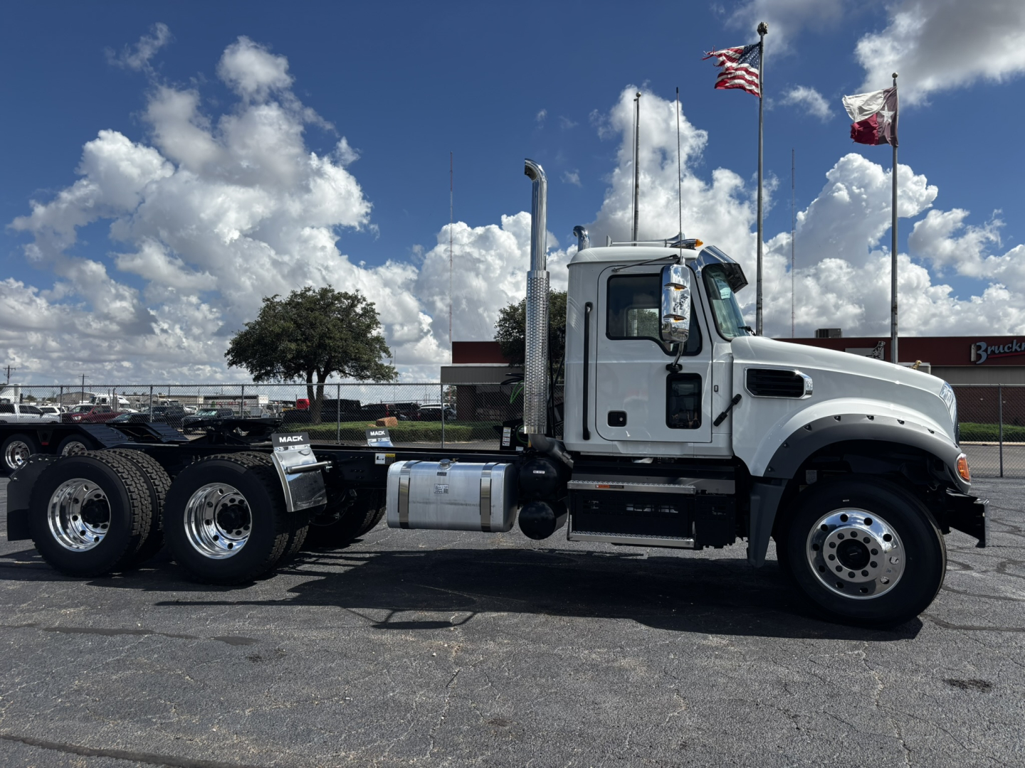 Side view of a white 2027 Mack GR64FT conventional daycab truck with dual rear axles, chrome wheels, and tall exhaust stacks; clear sky with flags in background.