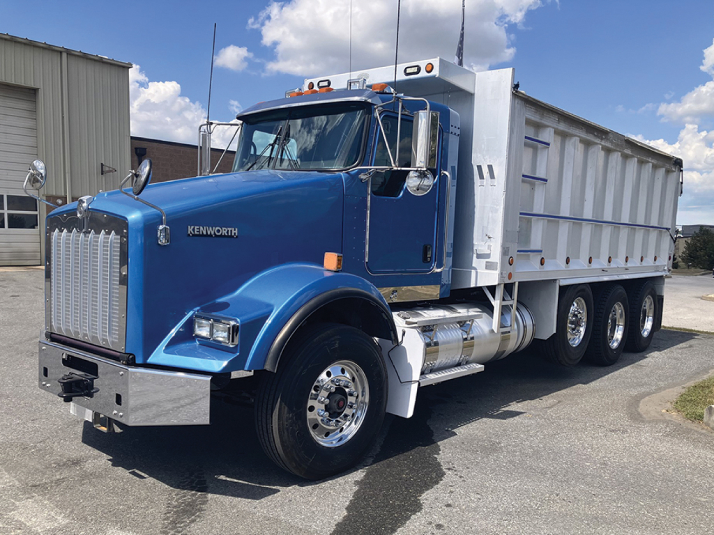 Kenworth T-800 dump truck in blue with chrome grille and silver dump bed, side view showing dual rear axles, chrome exhaust stacks, and Kenworth badge.