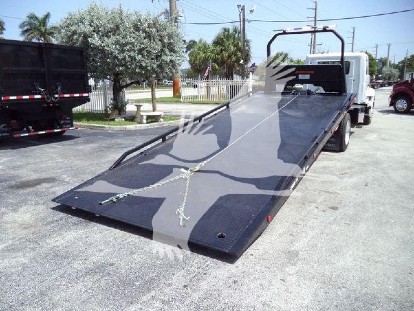 Truck with a lowered flatbed/ramp in a rollback configuration; white cab, black steel ramp surface, chains securing the platform, outdoor lot.