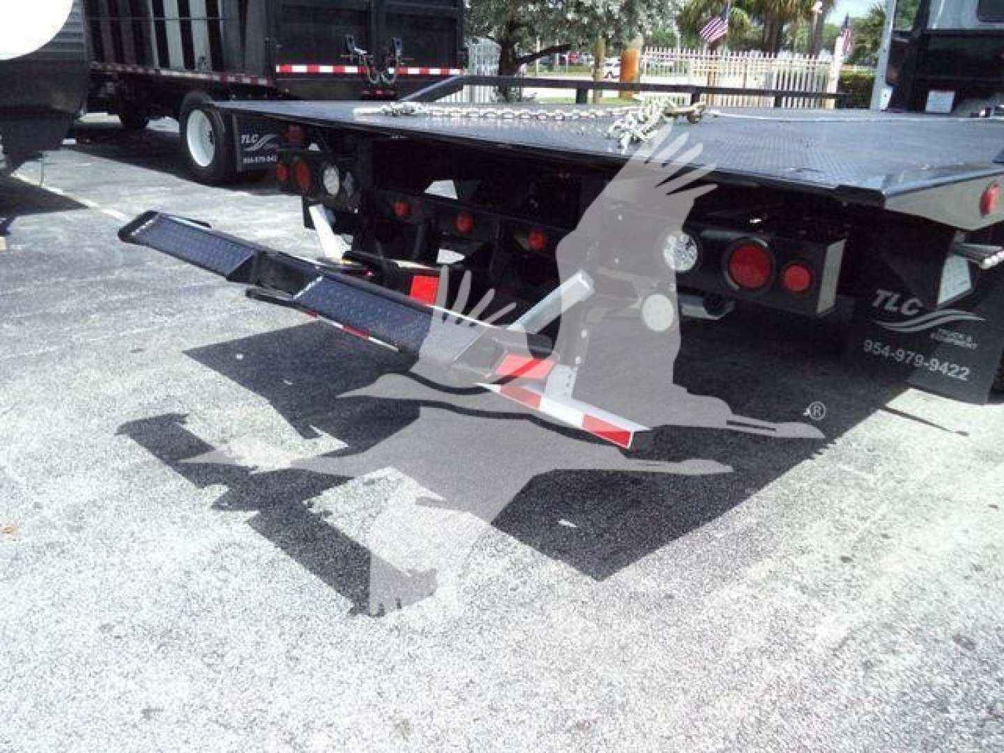Flatbed tow truck rear with hydraulic ramp and taillights; visible metal platform, safety lights, and ramps folded under the bed; black and white coloring visible.
