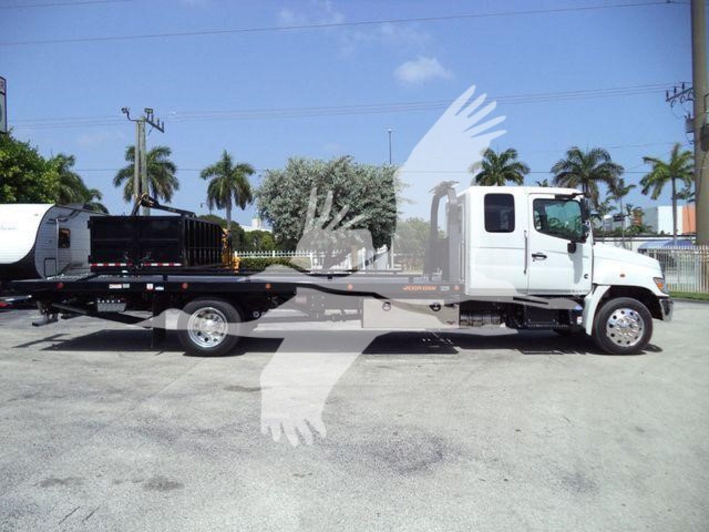 Truck with rollback bed and crane attachment on a lot, white cab, palm trees in background.
