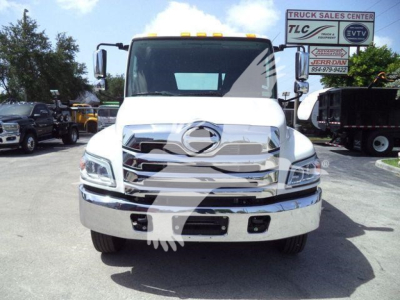 Front view of a white 2026 Hino L6 rollback truck showing chrome grille and bumper, with dealership lot in background.