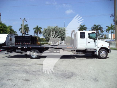 Truck with rollback bed and crane attachment on a lot, white cab, palm trees in background.