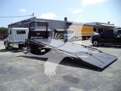 Front left view of a 2026 Hino L6 rollback truck with a long flatbed ramp extended, visible cab and rear truck in background.