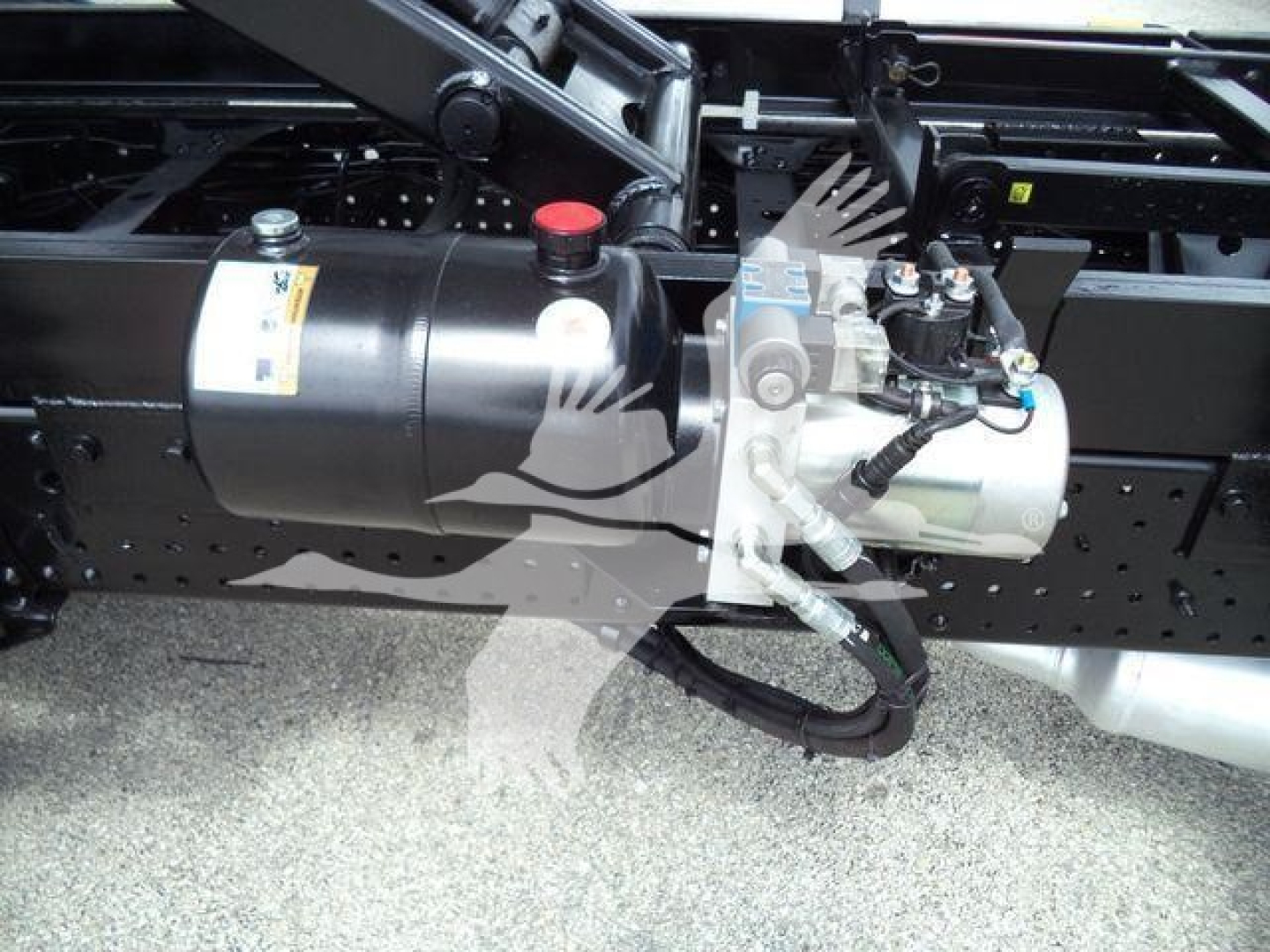 Truck chassis with fuel tank and auxiliary pump/valve assembly visible; includes black cylindrical tank with a red cap and a silver mechanical unit with hoses attached.