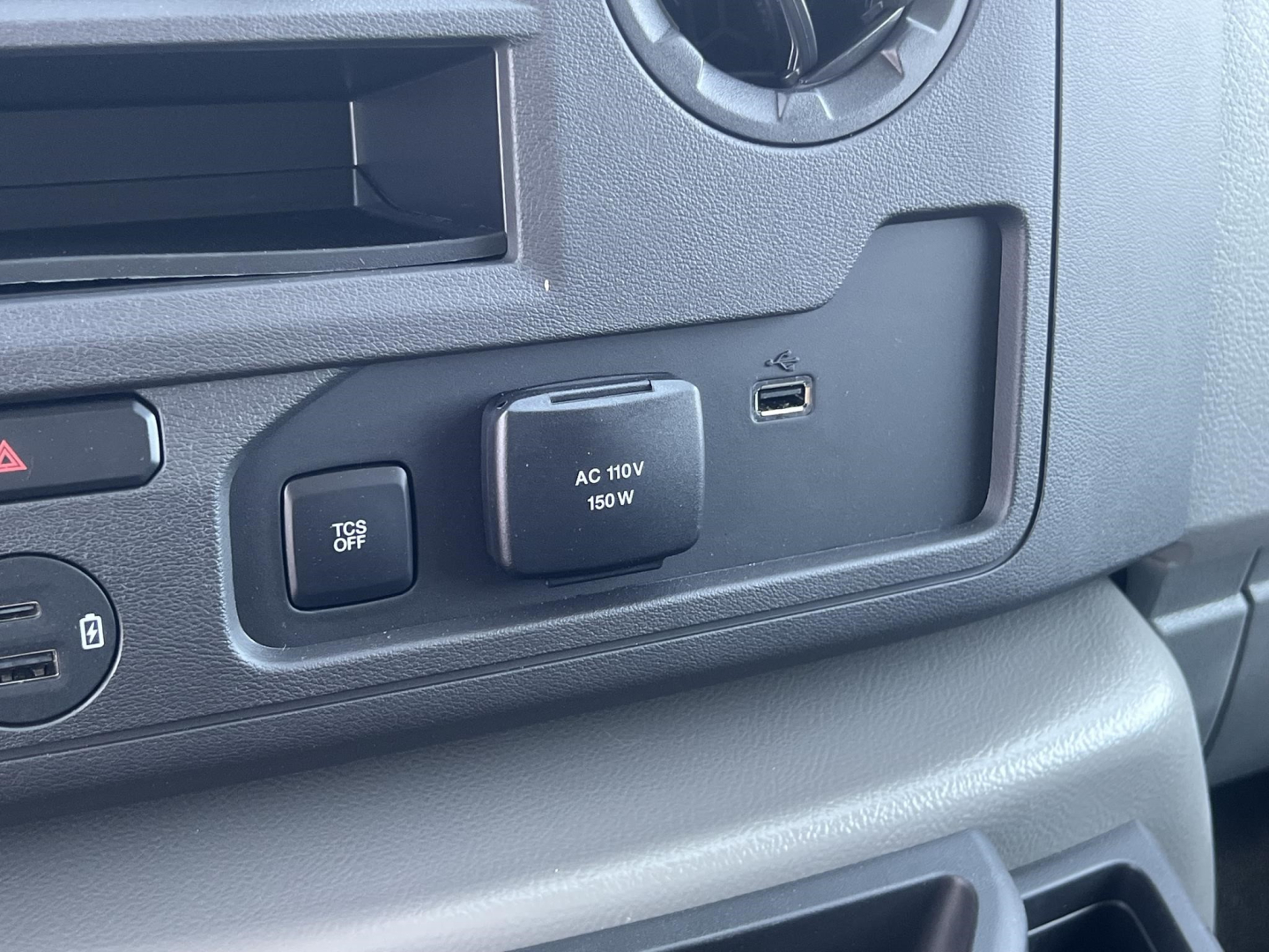 Ford E-Series cabin panel showing 12V/AC power outlet labeled AC 110V 150W and a USB-C port, plus a TCS OFF button and circular vent near the dashboard edge.