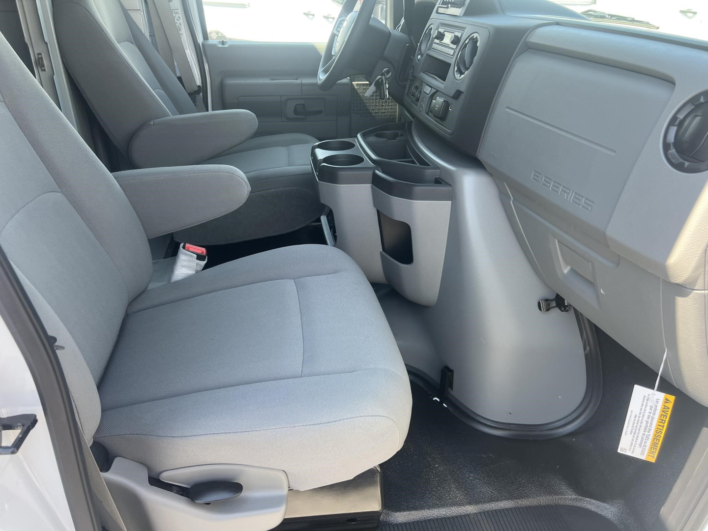 Ford E350 interior front cabin view showing driver and passenger seats, center console with cup holders, dashboard, steering wheel, and entry area.