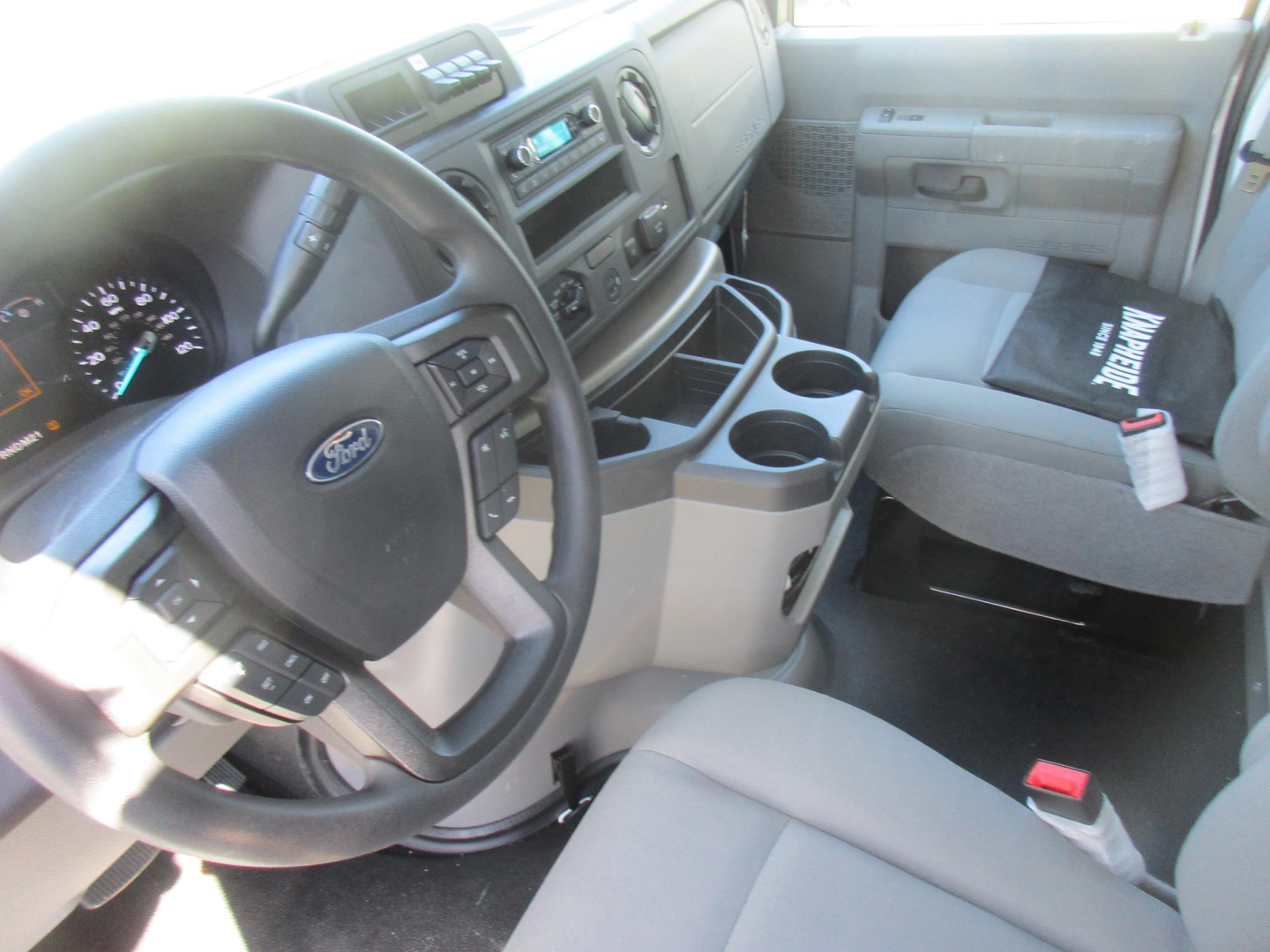 Ford E-350 cab interior showing steering wheel with Ford badge, dashboard controls, center console with cup holders, grey seats, and a black document or cover on the passenger seat.