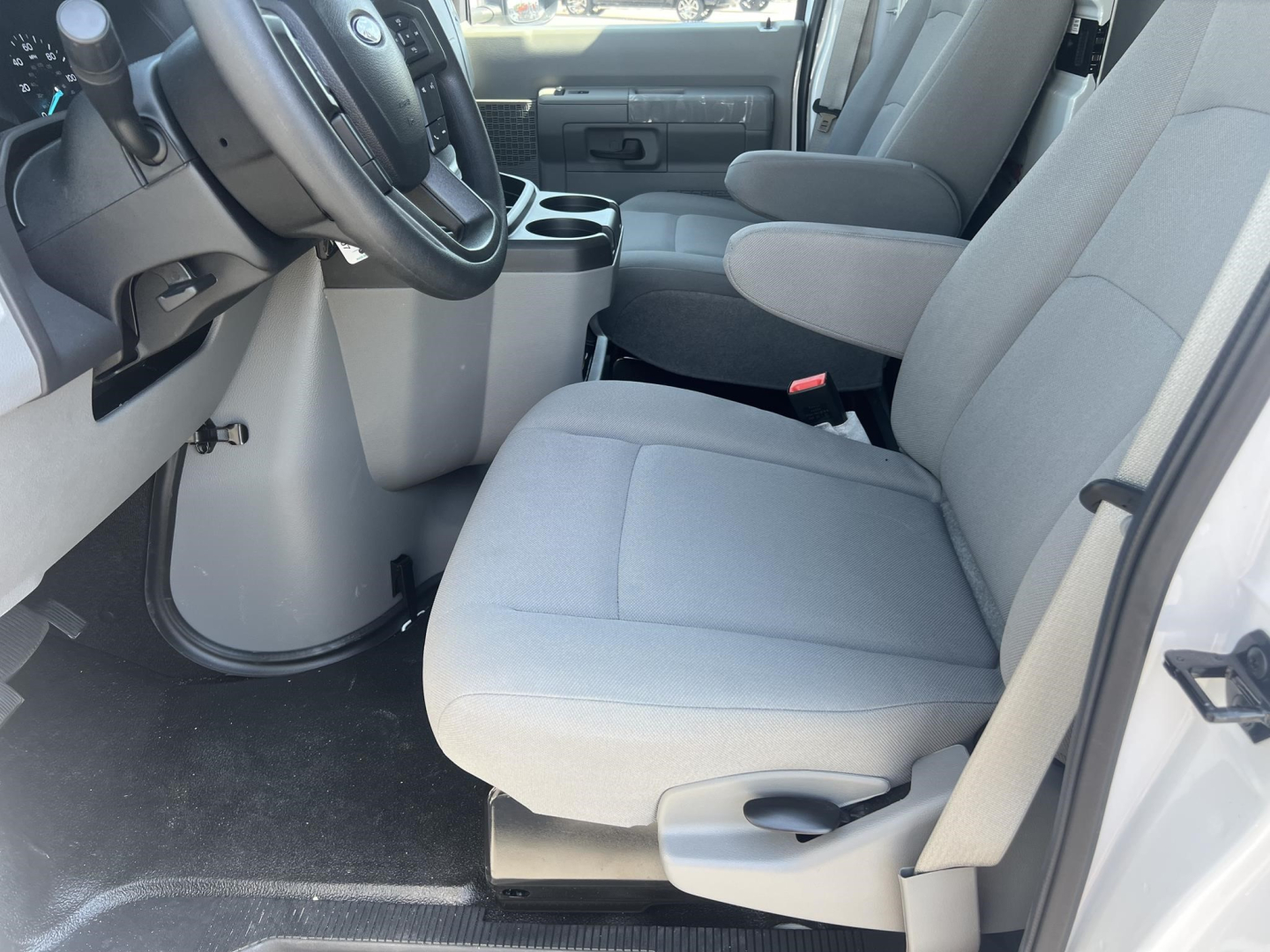 Front interior of a 2026 Ford E350 cargo van showing driver and passenger seats, center console, and steering wheel. Light gray fabric seats, gray door panels, and black floor.