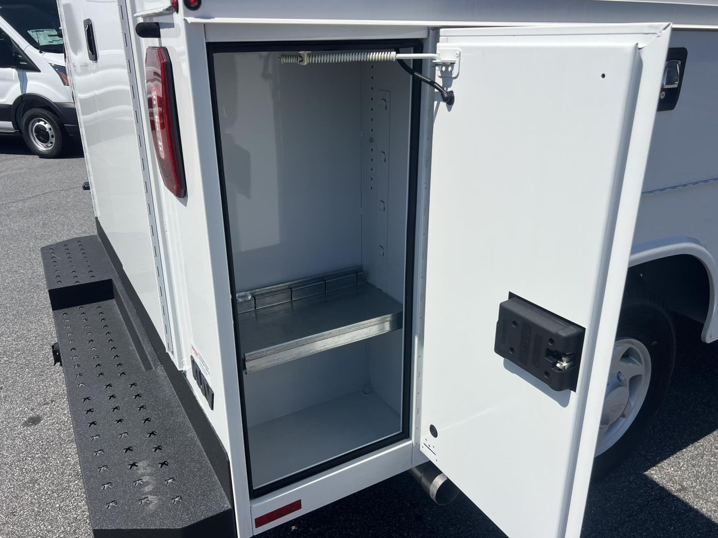 Truck cargo area of a white 2026 Ford E350 service/utility van showing an open rear cabinet with a lockable door, interior shelves, and step bumper.