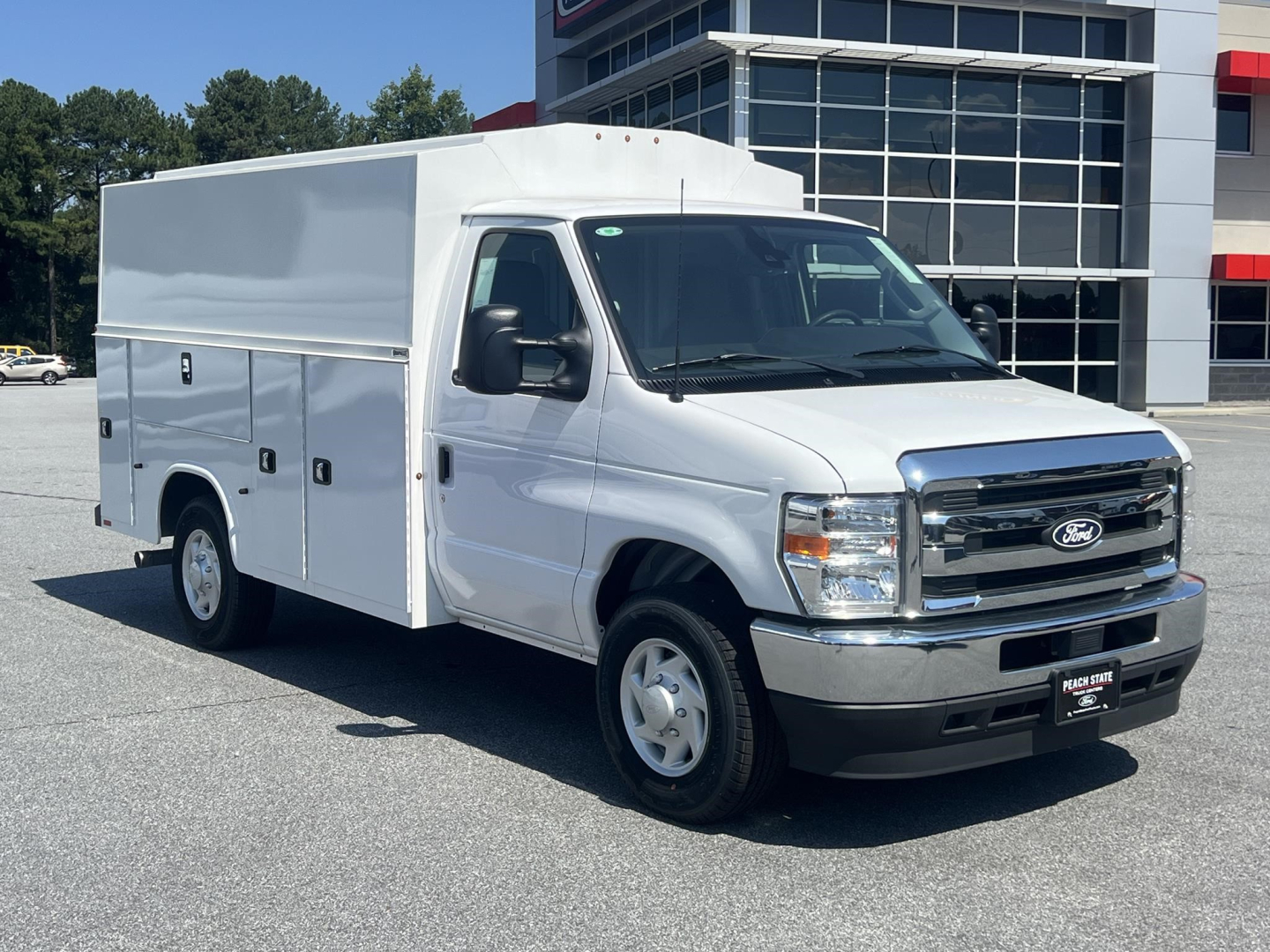 Ford E-350 service body van with utility compartments on side, front grille and headlights visible, white exterior, dealership lot in background.