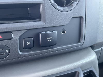 Ford E-Series cabin panel showing 12V/AC power outlet labeled AC 110V 150W and a USB-C port, plus a TCS OFF button and circular vent near the dashboard edge.