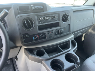 Ford E-Series Econoline interior showing dash with circular vents, factory stereo, climate controls, cup holders, and storage compartments in-front of passenger seat.