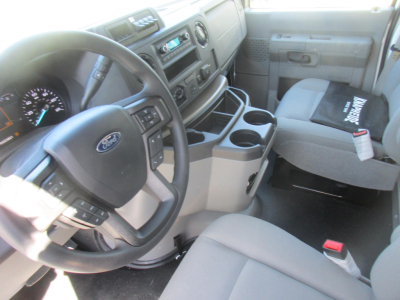 Ford E-350 cab interior showing steering wheel with Ford badge, dashboard controls, center console with cup holders, grey seats, and a black document or cover on the passenger seat.