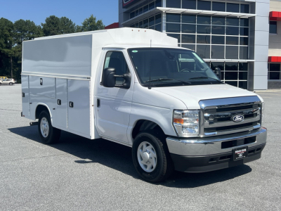 Ford E-350 service body van with utility compartments on side, front grille and headlights visible, white exterior, dealership lot in background.