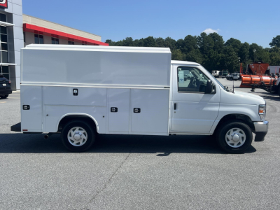 Side view of a white Ford E-350 service utility van with multiple external storage compartments along the side, cargo box on rear, standard dual rear wheels, and a single cab. No visible branding beyond generic Ford emblems.