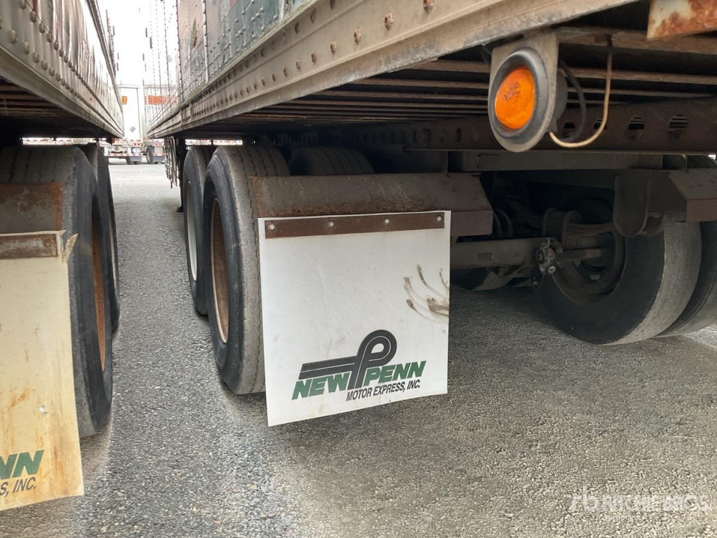 Trailer side view showing New Penn mudflap and dual wheels under trailer with orange side marker, metal frame, gravel ground.