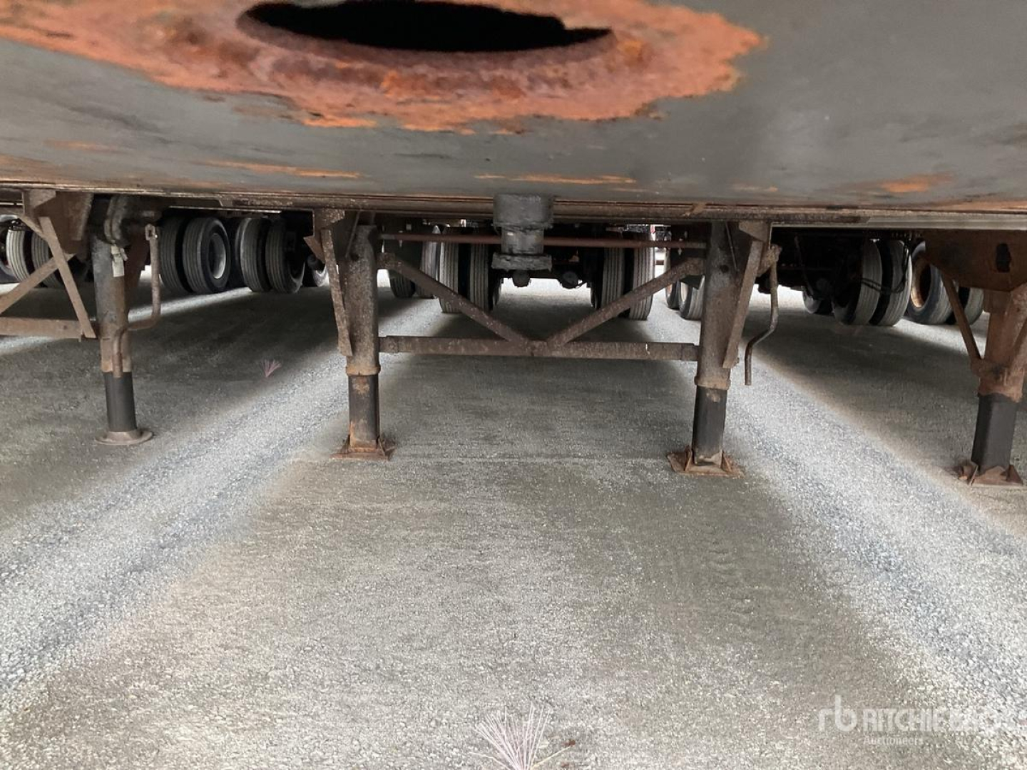 Underside of a trailer showing landing gear and suspension mounts with rust on the frame and rusty circular cutout in the floor, view from ground level under a trailer.
