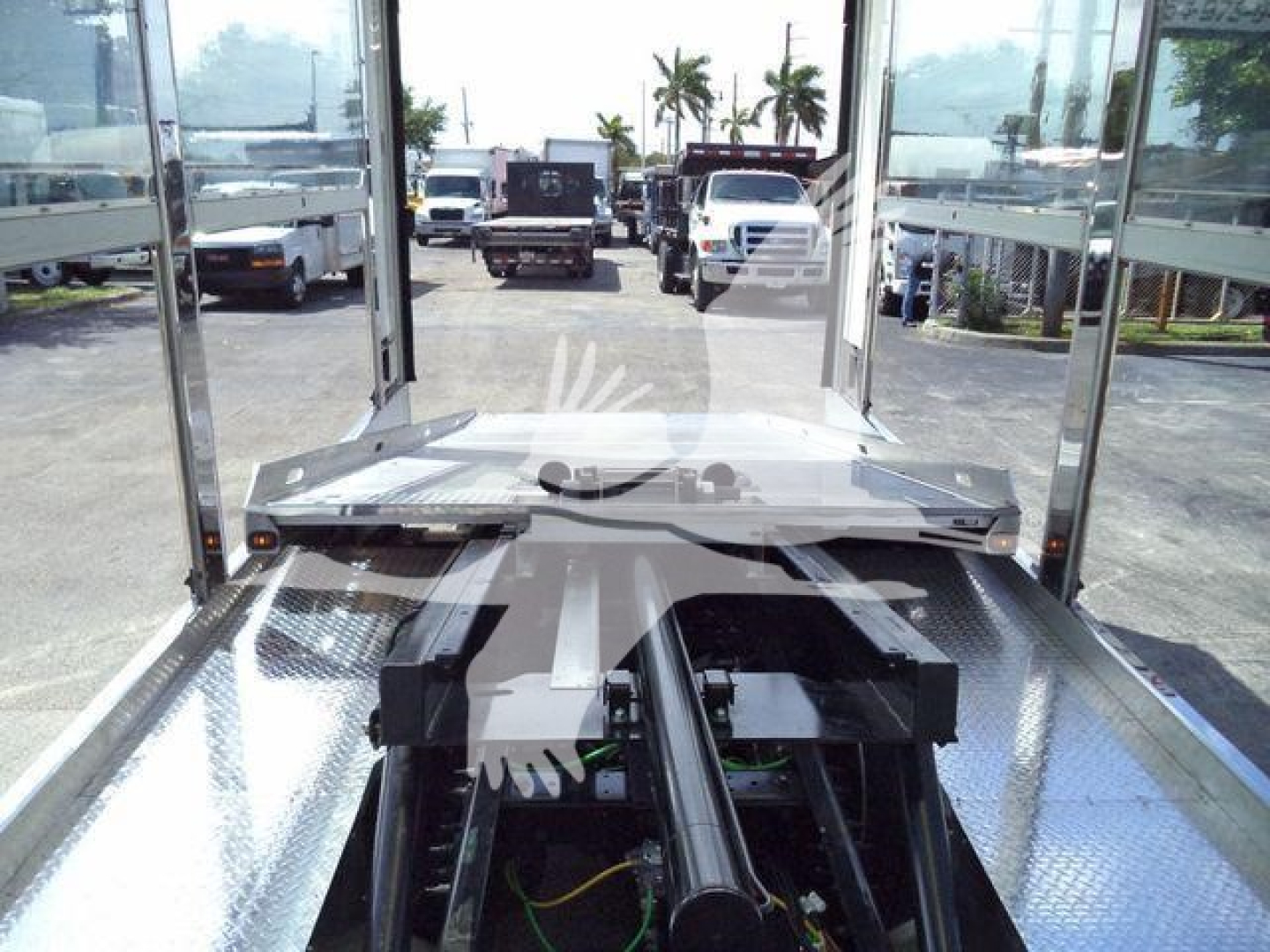 Truck rear view showing a tow truck bed with a black hydraulic piston and aluminum decking; surrounding chrome rails and a distant vehicle line.