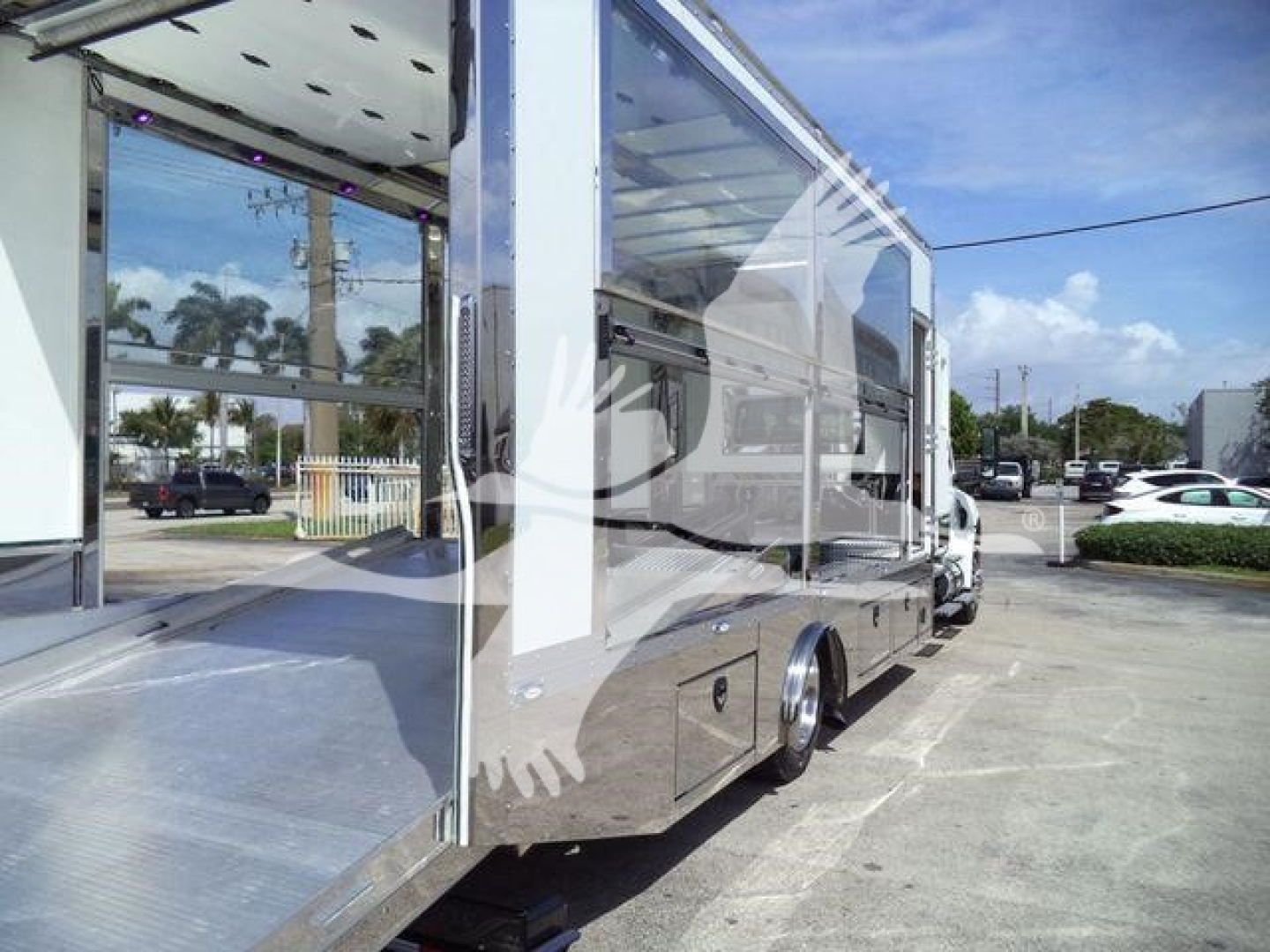 Truck or vehicle body in a showroom lot with side extension and reflective mirrored panels. Open side ramp/dock at left with metallic surface. Wheel and chrome trim visible.