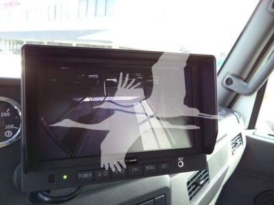 Truck cab dash with large touchscreen monitor screen showing a reflection of the interior; control buttons along bottom; power indicator light on left; dashboard and window visible.