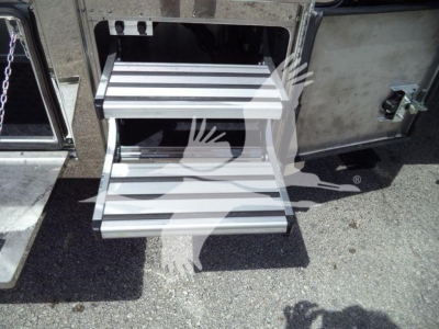 Two metal stairs and side of a truck with a chain and latch visible. Steps show ribbed aluminum treads and drop-down access panel.