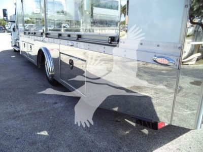 Side view of a new International MV rollback truck with polished metal body panels, dual rear wheels, storage compartments, and reflection on body from sun.