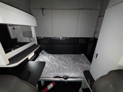 Kenworth T680 sleeper cab interior showing bed platform in cabin with storage cabinet on left and step on right, plastic covering on mattress.