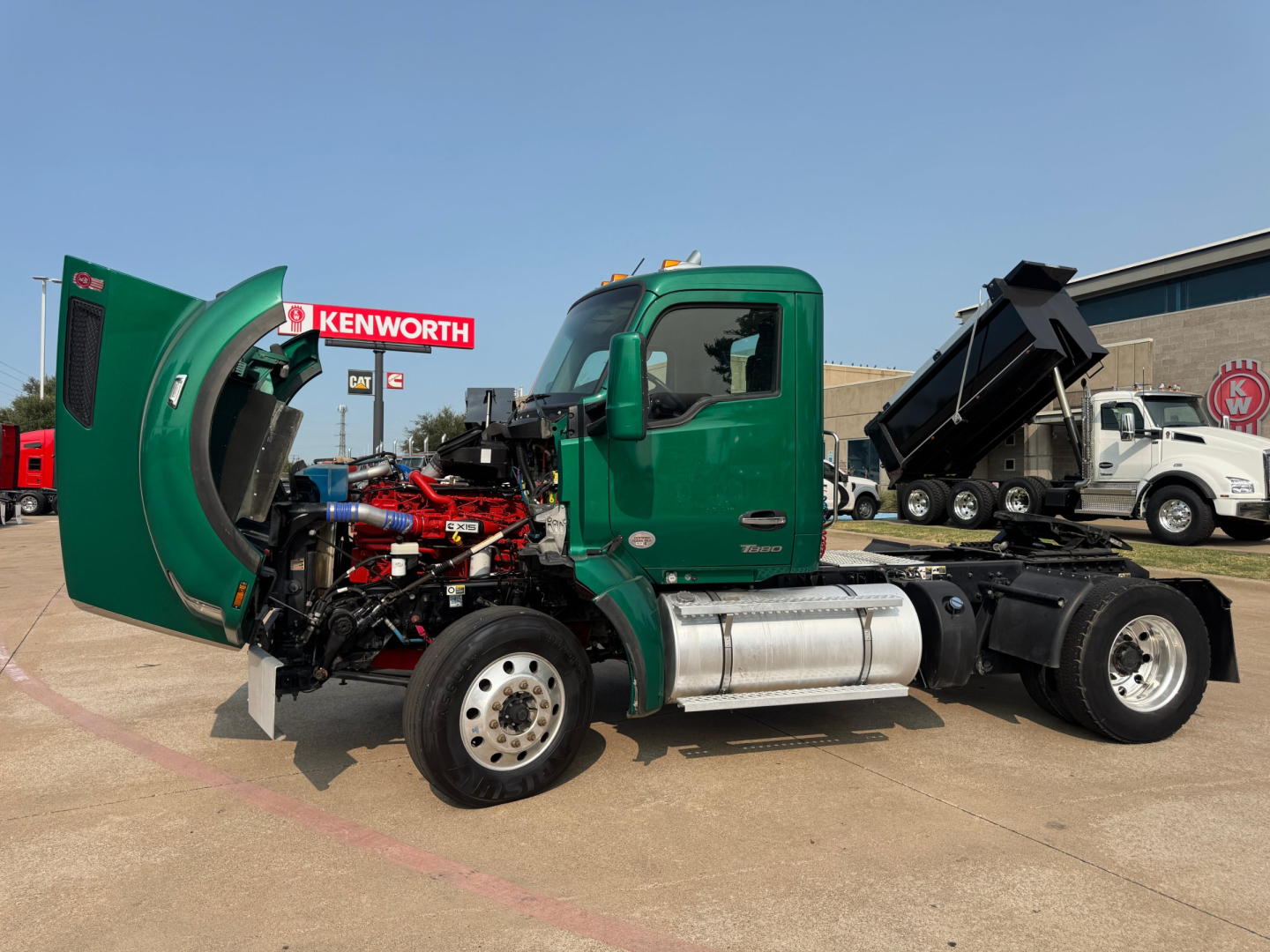 Kenworth T880 conventional daycab truck with hood open showing engine and red components; green cab and chassis; large fuel tanks; dump bed in back; dealership lot.