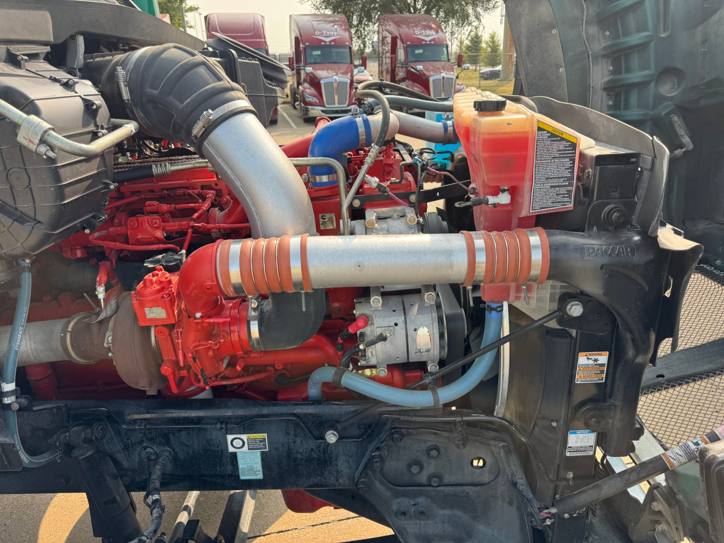 Engine bay of a 2022 Kenworth T880 with red engine components, blue hoses, orange coolant reservoir, and large intercooler piping visible.