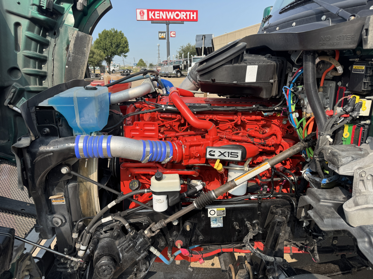 Kenworth T880 engine bay showing X15 engine with red block and blue hoses, various cables, and battery area visible.