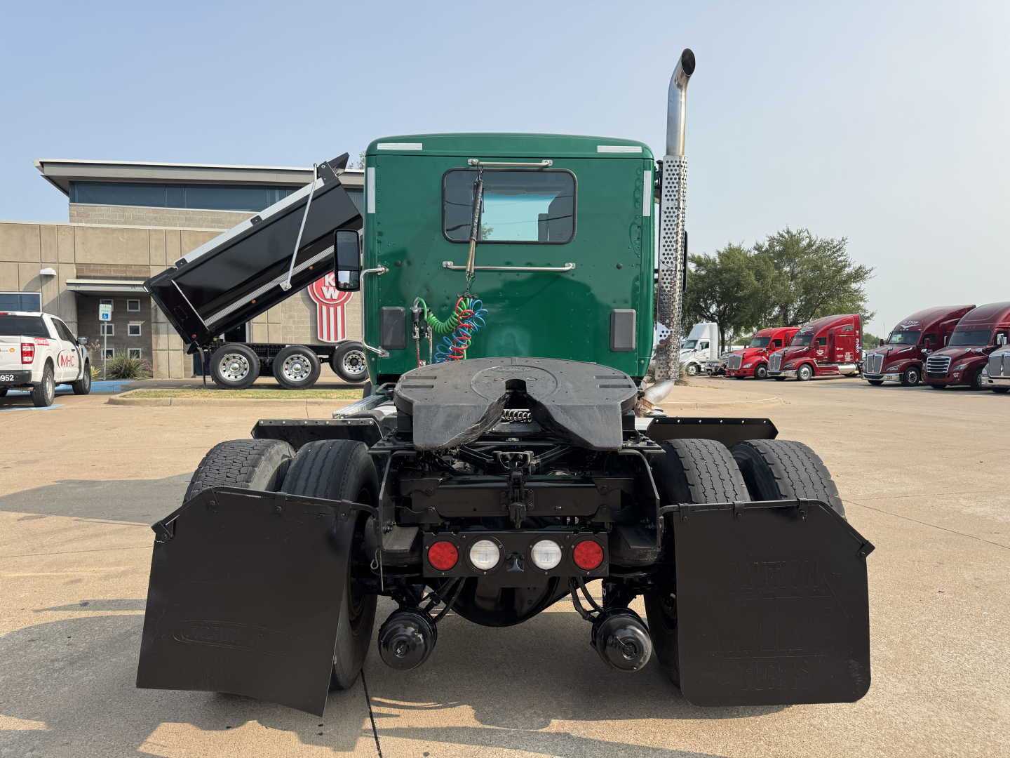Kenworth T880 rear view showing dual rear wheels, mud flaps, and exhaust stack with green cab in background.