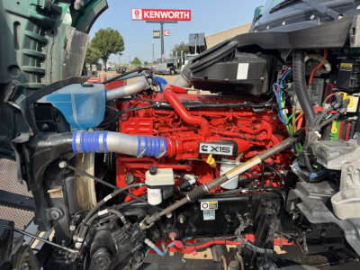 Kenworth T880 engine bay showing X15 engine with red block and blue hoses, various cables, and battery area visible.