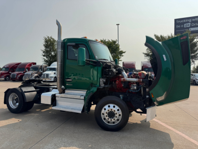 Kenworth T880 conventional daycab truck, green cab with hood open showing engine components, chrome exhaust stack, front wheel, side step, and raised hood on a dealership lot.