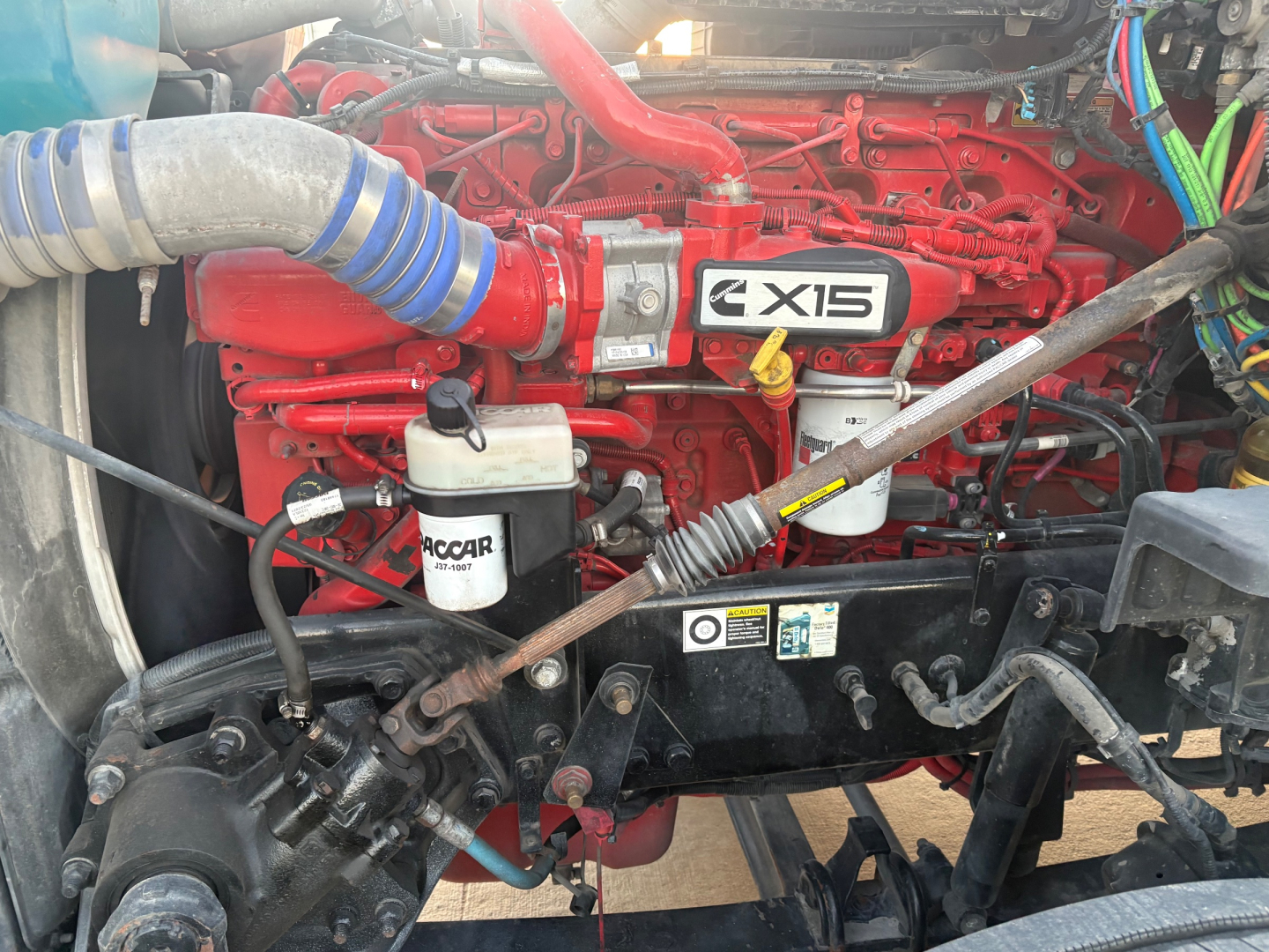 Kenworth T680 engine bay showing Cummins X15 badge and coolant reservoir with hoses and wiring harnesses.