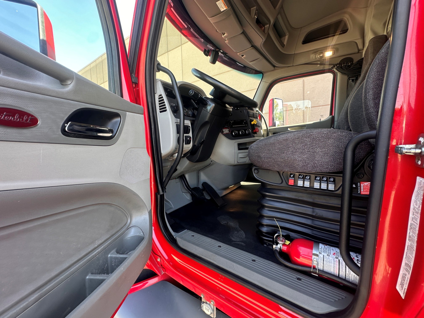 Peterbilt 579 cab interior with driver seat and steering wheel visible; door panel shows Peterbilt logo; open door reveals dashboard, controls, and a fire extinguisher in the cab floor area.